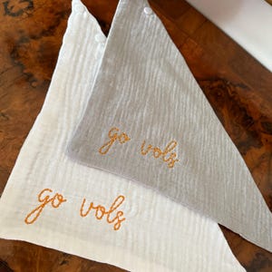 May include: Two triangular baby bibs, one white and one gray, with the embroidered text "go vols" in orange thread. The bibs are made of a textured fabric and have a button closure at the top.