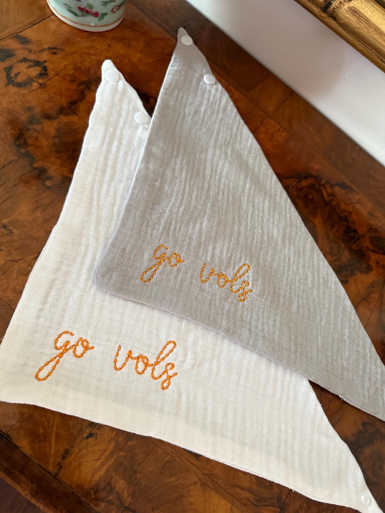 May include: Two triangular baby bibs, one white and one gray, with the embroidered text "go vols" in orange thread. The bibs are made of a textured fabric and have a button closure at the top.