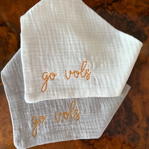 May include: Two triangular baby bibs, one white and one gray, are stacked on top of each other. The words "go vols" are embroidered in orange thread on each bib. The bibs are made of a soft, textured fabric.