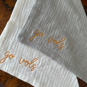 May include: Two cloth napkins, one white and one gray, are displayed. The phrase "Go Vols" is embroidered in orange thread on each napkin. The napkins are folded and appear to be made of a textured fabric.