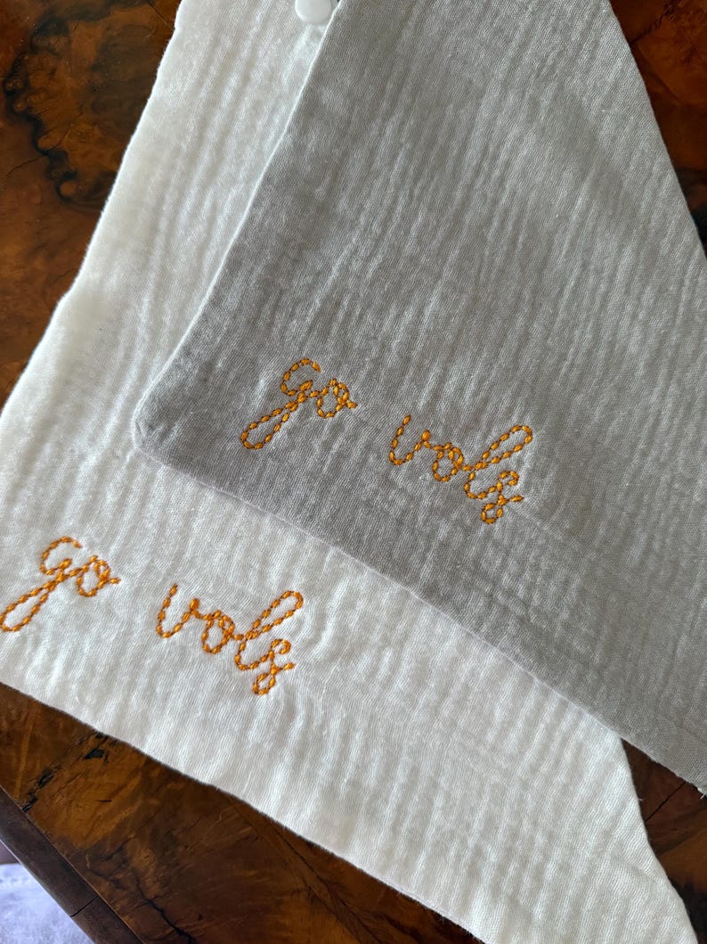 May include: Two cloth napkins, one white and one gray, are displayed. The phrase "Go Vols" is embroidered in orange thread on each napkin. The napkins are folded and appear to be made of a textured fabric.