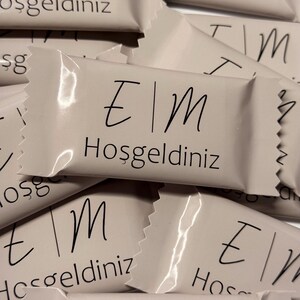 Könnte beinhalten: Eine Ansammlung kleiner, rechteckiger, hellfarbener Verpackungen. Jede Verpackung trägt die Buchstaben "E|M" und das Wort "Hoşgeldiniz" in schwarzer Schrift. Die Verpackungen sind leicht überlappend angeordnet.