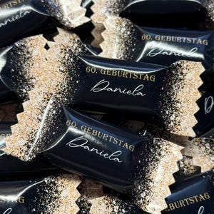 Könnte beinhalten: Schwarze und goldene Gastgeschenke mit dem Aufdruck "60. Geburtstag Daniela". Die Boxen sind in Form von Bonbonverpackungen und haben eine glitzernde Oberfläche.