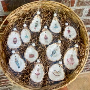 May include: A collection of oyster shell Christmas ornaments with gold trim and various festive illustrations. The ornaments feature images of a nutcracker, ballerina, and other holiday-themed figures. They are displayed in a woven basket.
