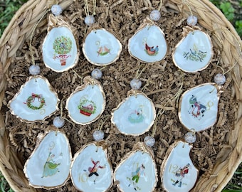 May include: A collection of oyster shell ornaments with gold trim, each featuring a unique hand-painted design. The ornaments depict various festive scenes, including birds, wreaths, and figures, with gold string hangers and beaded accents. They are arranged in a woven basket.