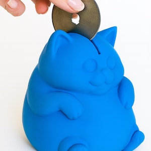 May include: A bright blue cat-shaped piggy bank with a coin slot on top. A hand is inserting a coin into the slot. The bank has a smiling face and rounded features. Two metal tools are on the white surface.