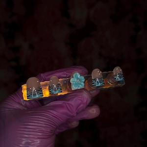 May include: A set of five fake nails with different designs. The nails are on a display card with a light blue flower design. The nails are in a hand wearing a purple glove.