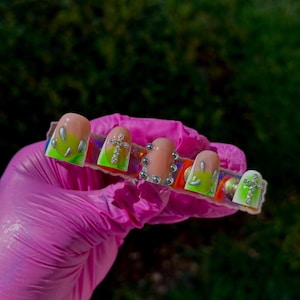 May include: A set of five artificial nails. The nails feature a gradient design with a nude base, transitioning to a neon green and silver accents. Each nail is adorned with silver embellishments, including crosses and rhinestones. The nails are held by a gloved hand.