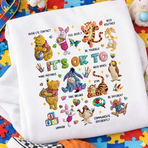 Puede incluir: Sudadera blanca con ilustraciones coloridas de personajes de Winnie the Pooh y frases como "It's OK to" y "Be Yourself." El diseño incluye el texto "Avoid Eye Contact," "Make Mistakes," y "Unmask."