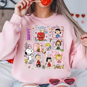 Puede incluir: Sudadera rosa pálido con temática de Pascua, con Snoopy y los personajes de Peanuts. El diseño incluye las palabras "Happy" y "Easter", con personajes con orejas de conejo, rodeados de huevos y flores de Pascua.