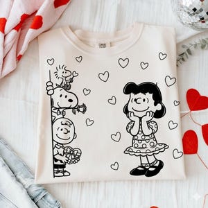 Puede incluir: Sudadera de color crema con un dibujo lineal negro de Snoopy, Woodstock, Charlie Brown y Lucy de Peanuts. El diseño incluye corazones y una caja de regalo. La sudadera tiene cuello redondo.