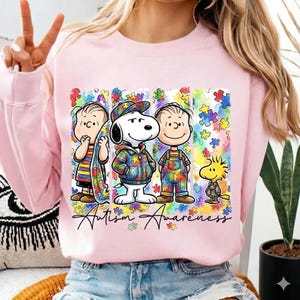 Puede incluir: Sudadera rosa claro con un gráfico colorido de Snoopy y sus amigos con patrones de piezas de rompecabezas. El texto "Autism Awareness" está escrito debajo. Los personajes visten trajes con diseños de piezas de rompecabezas.