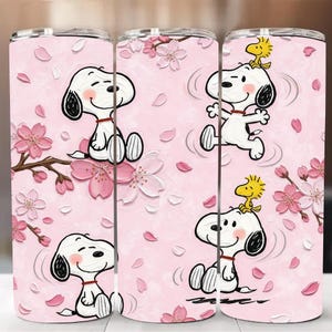 Puede incluir: Un vaso rosa con diseños de Snoopy y Woodstock, con flores de cerezo. El vaso tiene una apariencia de acero inoxidable en la parte superior e inferior. El diseño incluye a Snoopy en varias poses, rodeado de flores y pétalos rosas.