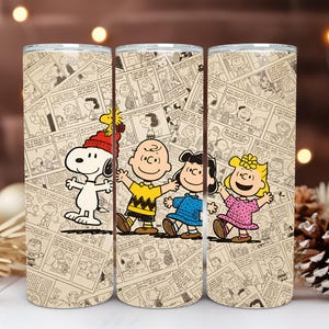 May include: Three stainless steel tumblers featuring the Peanuts gang. Each tumbler has a beige background with comic strip panels. Snoopy wears a red hat, Charlie Brown has a yellow shirt, Lucy wears blue, and Sally wears pink.