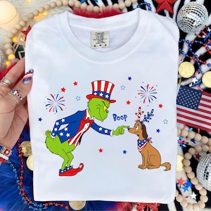 Puede incluir: Una camiseta blanca con un gráfico de un personaje verde con sombrero de copa y traje rojo, blanco y azul, tocando la nariz de un perro marrón con pañuelo patriótico y diadema de astas. El texto Boop está impreso en color azul.