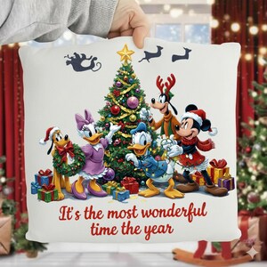 Puede incluir: Cojín cuadrado blanco con un árbol de Navidad y personajes de Disney, incluyendo Mickey, Minnie, Donald, Daisy, Pluto y Goofy. El cojín tiene el texto "It's the most wonderful time the year" en rojo.