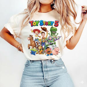 May include: A cream-colored short-sleeve t-shirt featuring a colorful graphic print of Toy Story characters including Woody, Buzz Lightyear, Jessie, Rex, Bullseye, Slinky Dog, Mr. Potato Head, Hamm, and an alien with the text Toy Story 5 above.
