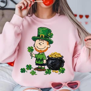 Puede incluir: Sudadera rosa claro con un personaje de dibujos animados disfrazado de duende, sosteniendo un trébol de cuatro hojas y una olla de oro. El duende lleva un sombrero y un atuendo verdes. Varios tréboles verdes están dispersos.