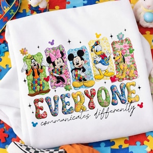 Puede incluir: Sudadera blanca con los personajes de Disney Goofy, Minnie Mouse, Mickey Mouse, Pato Donald y Pluto. La palabra "EVERYONE" está en letras florales, con la frase "communicates differently" debajo. Los personajes y las letras están delineados con un patrón de rompecabezas colorido.
