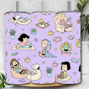 Puede incluir: Un juego de tres vasos de acero inoxidable con un fondo lavanda y un patrón de dibujos animados repetido. El patrón presenta a Snoopy, Charlie Brown, Lucy y otros personajes de Peanuts en varias poses relajantes, junto con estrellas, plantas y tazas de café.