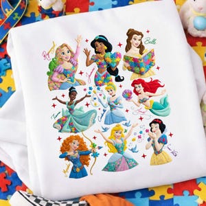 Puede incluir: Sudadera blanca con ilustraciones coloridas de princesas de Disney, incluyendo a Rapunzel, Jasmine, Bella, Ariel y Blancanieves. Cada princesa está representada con un diseño único, con sus nombres escritos en cursiva.