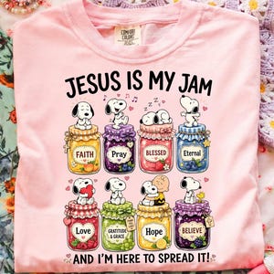 May include: A light pink t-shirt featuring a graphic of eight colourful jam jars topped with Snoopy characters. The text reads "JESUS IS MY JAM" at the top and "AND I'M HERE TO SPREAD IT!" at the bottom. Each jar is labelled with words like Faith, Pray, Blessed, Eternal, Love, Gratitude & Grace, Hope, and Believe.
