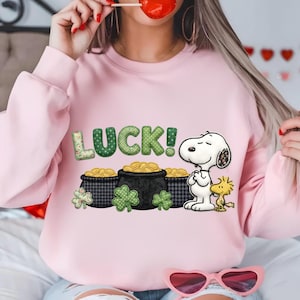 Puede incluir: Sudadera rosa claro con un diseño del Día de San Patricio. El gráfico presenta a Snoopy, Woodstock, ollas de oro y la palabra "LUCK!" en letras verdes. La letra "L" tiene un estampado floral.