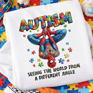 Puede incluir: Sudadera blanca con un diseño colorido de Spider-Man con piezas de rompecabezas. La palabra "AUTISMO" está escrita con letras de rompecabezas. El texto "SEEING THE WORLD FROM A DIFFERENT ANGLE" está impreso debajo.