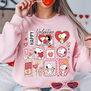 Puede incluir: Sudadera rosa claro con varios diseños de Snoopy y Woodstock para el Día de San Valentín. El diseño incluye corazones, la palabra "Valentine" y la palabra "Happy". La sudadera tiene cuello redondo y mangas largas.