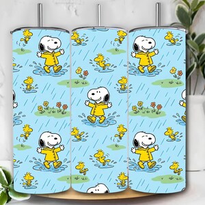 Puede incluir: Un vaso de acero inoxidable con un patrón repetido de Snoopy con un impermeable amarillo, chapoteando en charcos con Woodstock, sobre un fondo azul claro con lluvia y flores. El vaso tiene una tapa y una pajita plateadas.