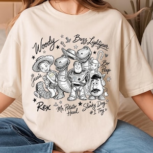 Puede incluir: Una camiseta de color crema con un gráfico de líneas negras de los personajes de Toy Story: Woody, Buzz Lightyear, Jessie, Rex, Mr. Potato Head, Bo Peep y Slinky Dog, con sus nombres en cursiva y el texto #1 Toy.