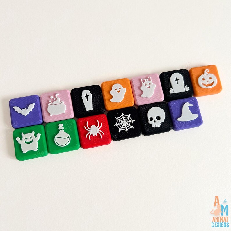 May include: A collection of fourteen square, multi-colored plastic tiles featuring raised white icons including a bat, cauldron, coffin, ghost, spider, web, skull, witch hat, and pumpkin. The bottom right corner displays the text Animai Designs.