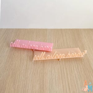 K&ouml;nnte beinhalten: Zwei rechteckige Schilder aus Acryl in Pastellfarben mit dem kursiven Text "fall asleep here". Ein Schild ist rosa, das andere pfirsichfarben. Die Schilder liegen auf einer hellen Holzoberfl&auml;che.