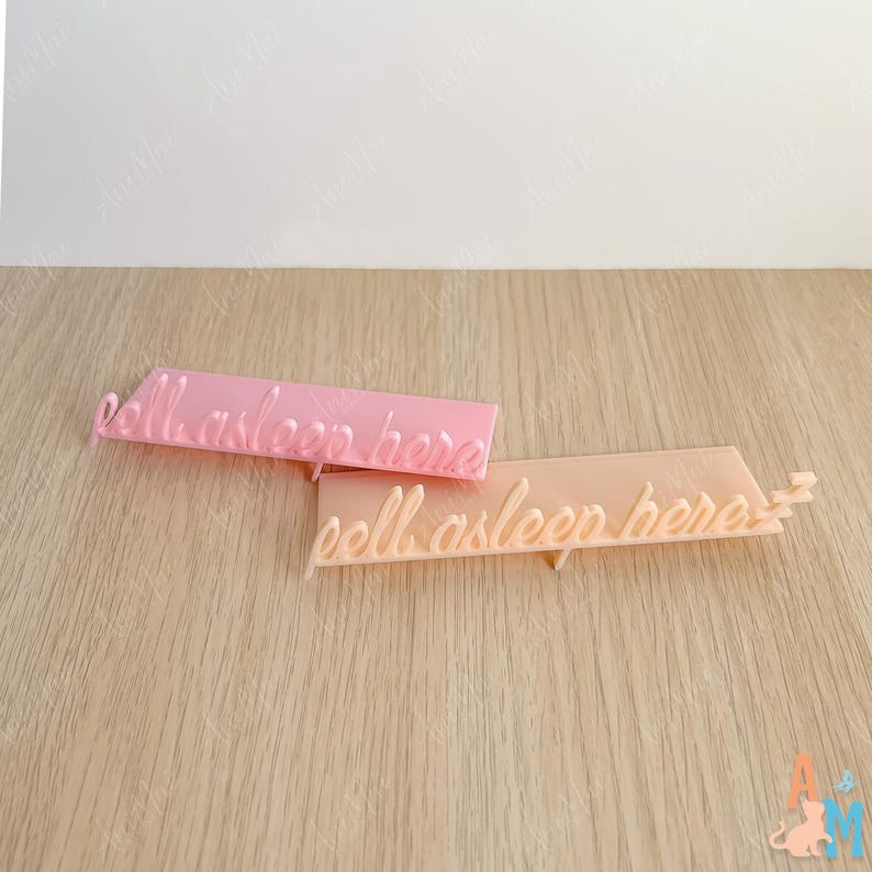 K&ouml;nnte beinhalten: Zwei rechteckige Schilder aus Acryl in Pastellfarben mit dem kursiven Text "fall asleep here". Ein Schild ist rosa, das andere pfirsichfarben. Die Schilder liegen auf einer hellen Holzoberfl&auml;che.