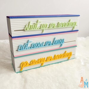 May include: A decorative book-shaped sign with the phrases "shut up, I'm reading", "not now, I'm busy", and "go away, I'm reading" in pastel colours. The sign has a white, textured surface and colourful accents.