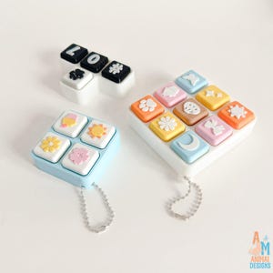 Op de afbeelding: Drie vierkante plastic bakjes met verhoogde iconen op de deksels. Een kleine zwart-witte set bevat letters en een pootafdruk. Twee grotere pastelkleurige sets bevatten iconen van bloemen, zonnen, manen, vlinders en strikjes met kettingen.