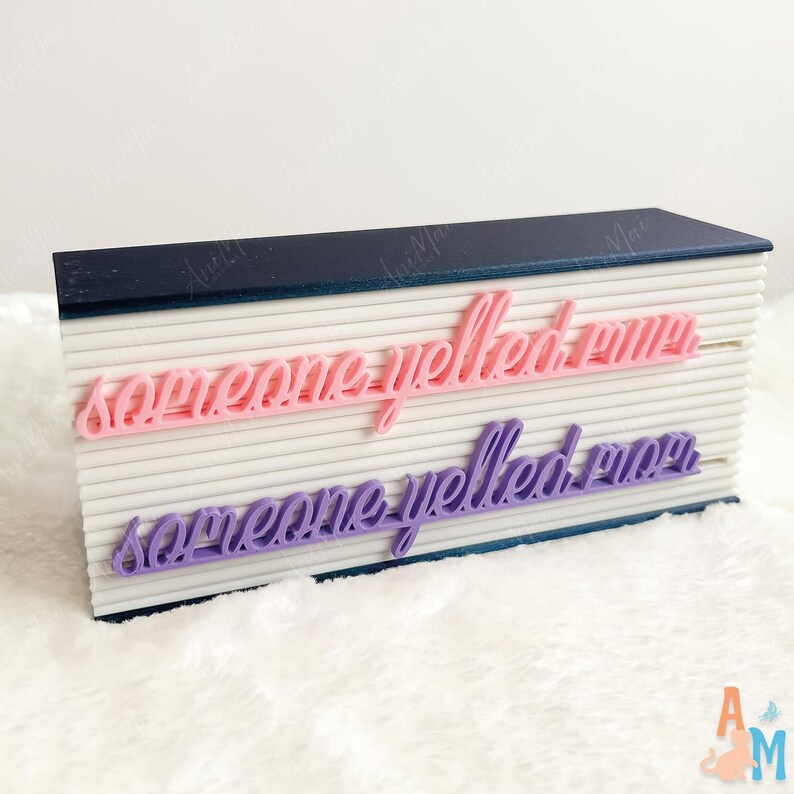 May include: A white, rectangular block with a navy blue top, featuring the words "someone yelled mom" in pink and purple script. The block has a ribbed texture.