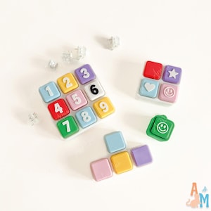 May include: A set of colorful square buttons featuring numbers one through nine, a red square, a purple star, a light blue heart, and two green and pink smiley faces. Several loose square buttons in blue, yellow, pink, and purple are included.