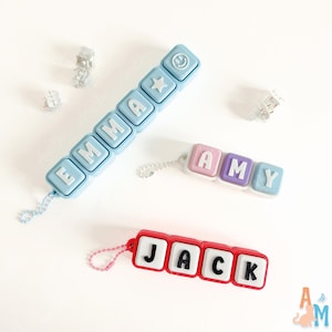 May include: Three custom name keychains made of square plastic blocks. One light blue keychain spells EMMA with a star and smiley face. One pink, purple, and blue keychain spells AMY. One red keychain spells JACK with white blocks and black letters.