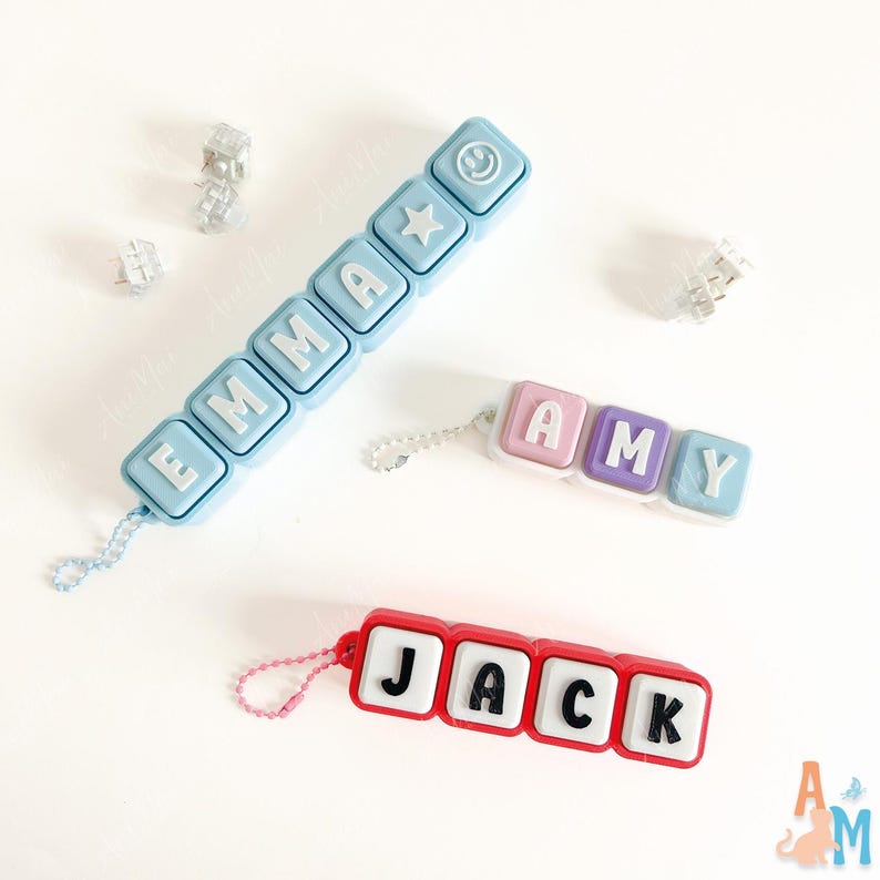 May include: Three custom name keychains made of square plastic blocks. One light blue keychain spells EMMA with a star and smiley face. One pink, purple, and blue keychain spells AMY. One red keychain spells JACK with white blocks and black letters.