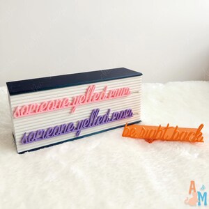 May include: A decorative sign with the text "someone yelled run" in pink and purple, and "be right back" in orange. The sign has a navy blue top and a white base with horizontal lines.