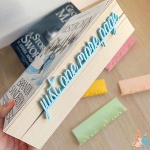 K&ouml;nnte beinhalten: Ein hellblaues Lesezeichen mit dem Text "just one more page" liegt auf einem Buch. Das Buch hat einen wei&szlig;en Einband und der Titel "Mistborn" ist sichtbar. Mehrere bunte Lesezeichen liegen um das Buch herum.