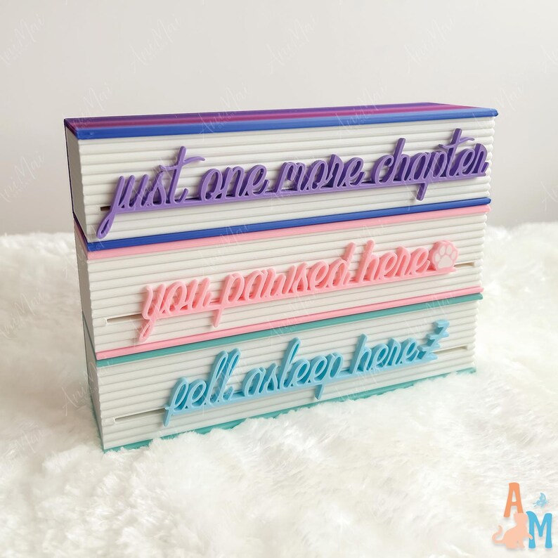 May include: A decorative book-shaped box with the phrases "just one more chapter", "you parked here", and "fell asleep here" in script lettering. The box has a white body with coloured accents in purple, pink, and blue.