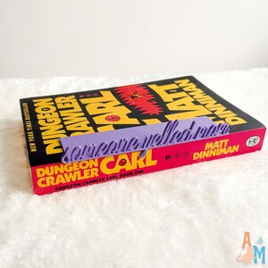 May include: A book titled "Dungeon Crawler Carl" with a purple bookmark that reads "someone yelled mom". The book has a red and yellow cover with the author's name, Matt Dinniman, and the words "New York Times Bestseller".