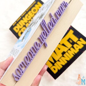 May include: A lilac bookmark with the words "someone yelled mom" in cursive is inserted into a book. The book has a pale brown cover and is held open. Two other books are visible in the background.
