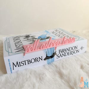 K&ouml;nnte beinhalten: Ein rosa Lesezeichen mit dem Schriftzug "fell asleep here" in Schreibschrift liegt auf einem Exemplar von "Mistborn: Book One" von Brandon Sanderson. Das Buch hat einen wei&szlig;en Einband mit blauen und schwarzen Illustrationen.