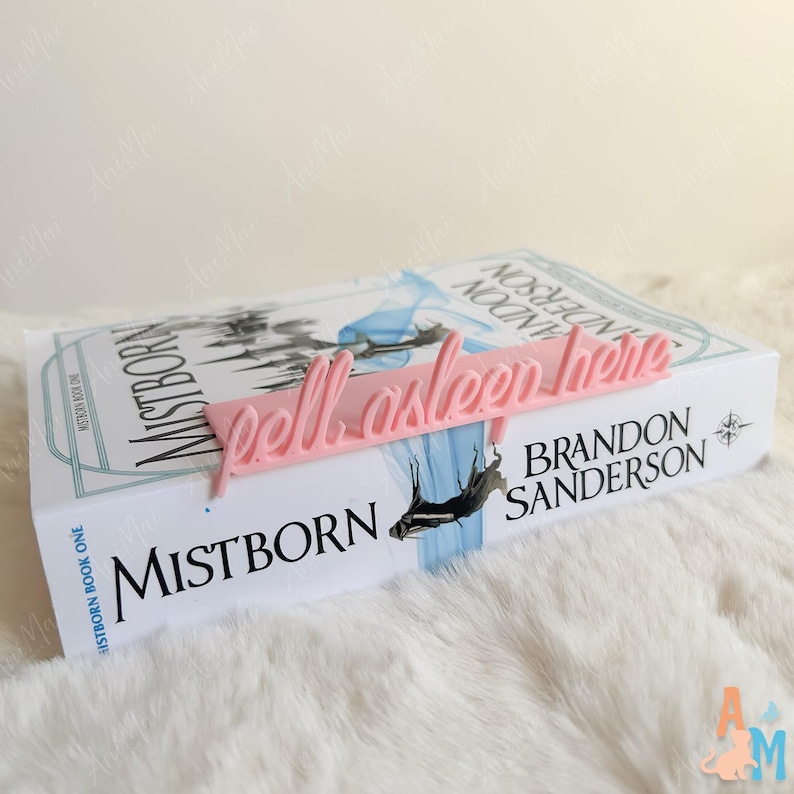 K&ouml;nnte beinhalten: Ein rosa Lesezeichen mit dem Schriftzug "fell asleep here" in Schreibschrift liegt auf einem Exemplar von "Mistborn: Book One" von Brandon Sanderson. Das Buch hat einen wei&szlig;en Einband mit blauen und schwarzen Illustrationen.
