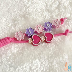 Puede incluir: Un accesorio de cord&oacute;n rosa con dos hilos distintos. Un hilo muestra cuentas en forma de flor en rosa claro, morado y rosa, deletreando el nombre SINDY. El segundo hilo presenta dos cuentas en forma de coraz&oacute;n en rosa oscuro con borde blanco.