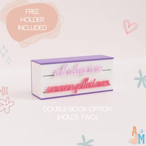 May include: A double book-shaped holder with a purple top and bottom and white pages. The holder has two lines of text: "fell asleep here" and "someone yelled mom." The text "FREE HOLDER INCLUDED" and "DOUBLE BOOK OPTION (HOLDS TWO)" are also visible.