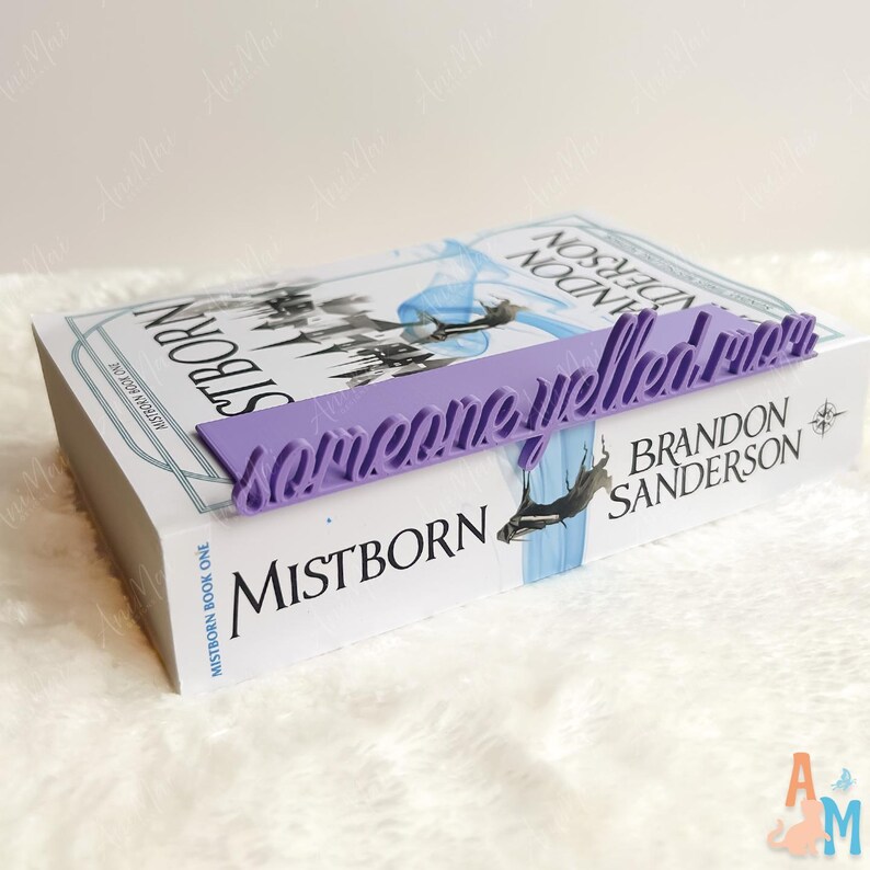 May include: A lilac bookmark with the words "Somewhere Welled Rain" rests on a book titled "Mistborn" by Brandon Sanderson. The book has a white cover with a blue and black illustration.